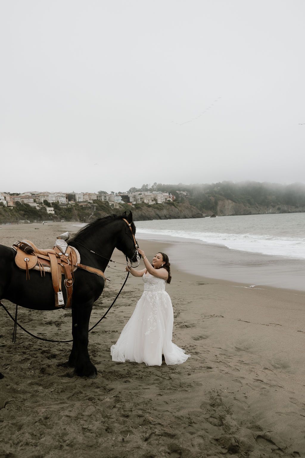 Photographer crouches to capture bride's wedding dress flowing in the wind on a misty beach landscape.