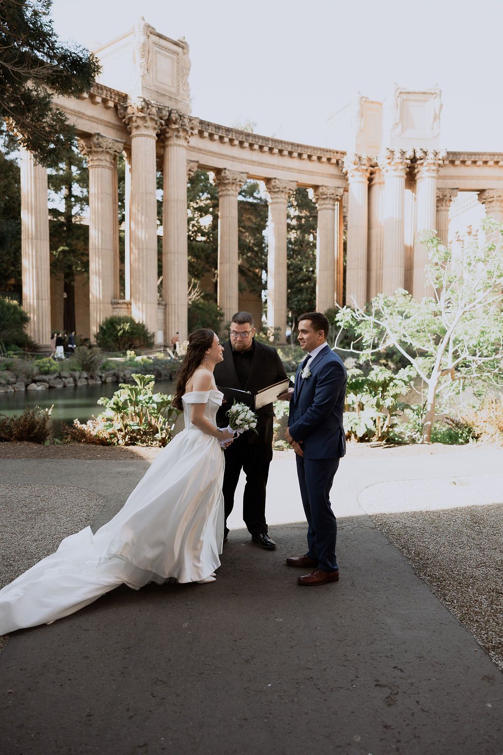 A wedding ceremony takes place under elegant stone columns in a classical garden setting.