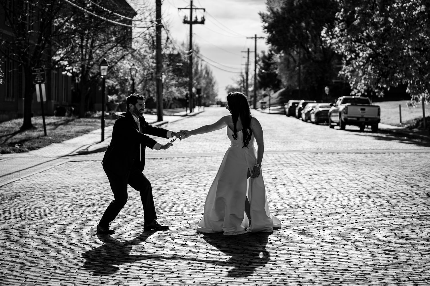 A couple in wedding attire dances playfully on a cobblestone street lined with parked cars.