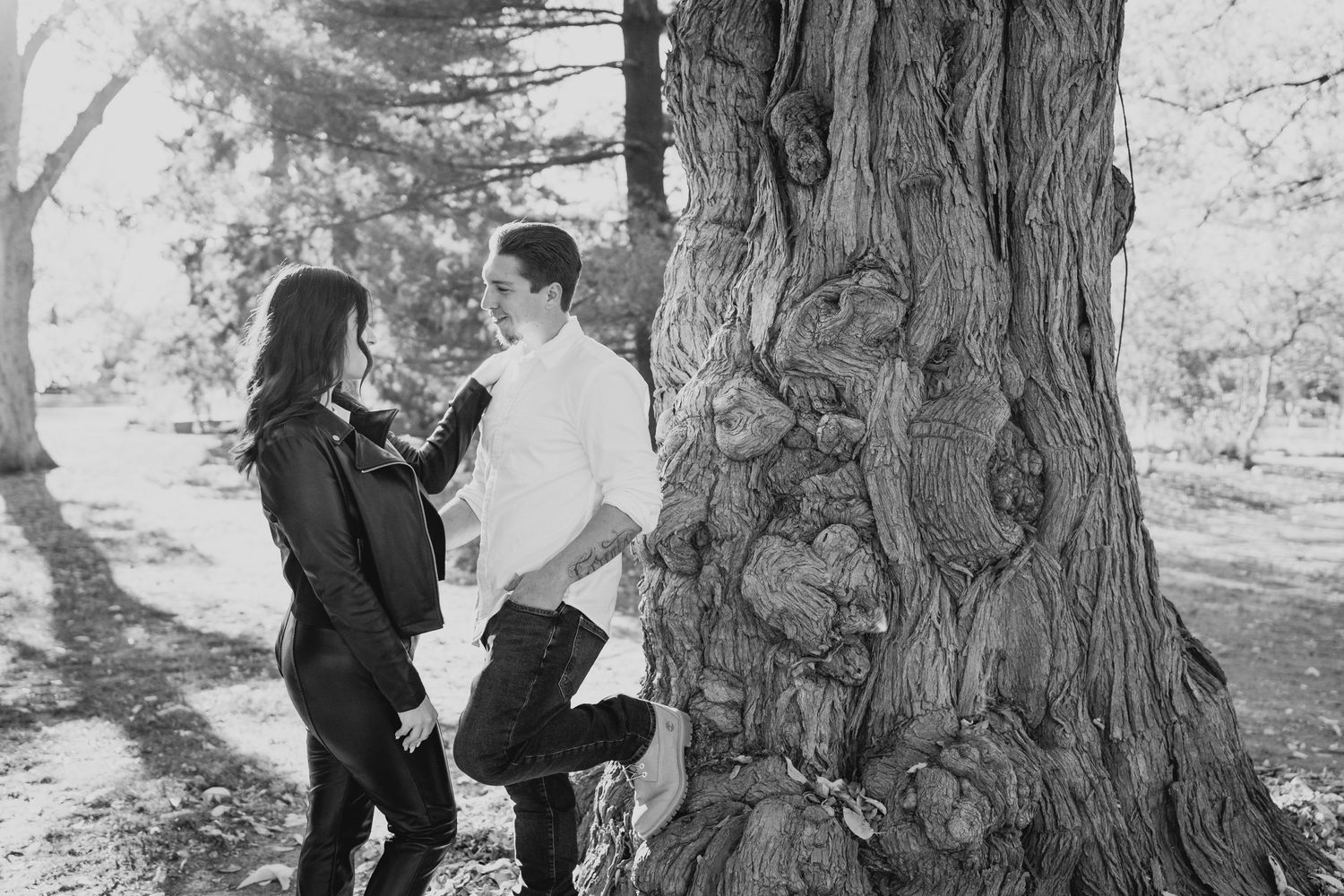 A couple poses intimately against a massive tree trunk in a black and white photograph.