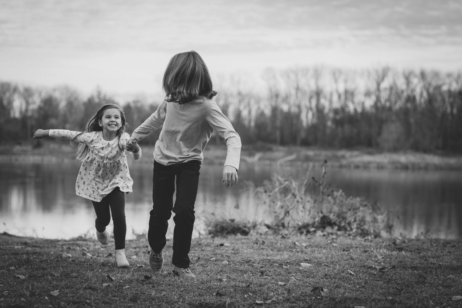 Two children run playfully alongside a lake during an overcast day.