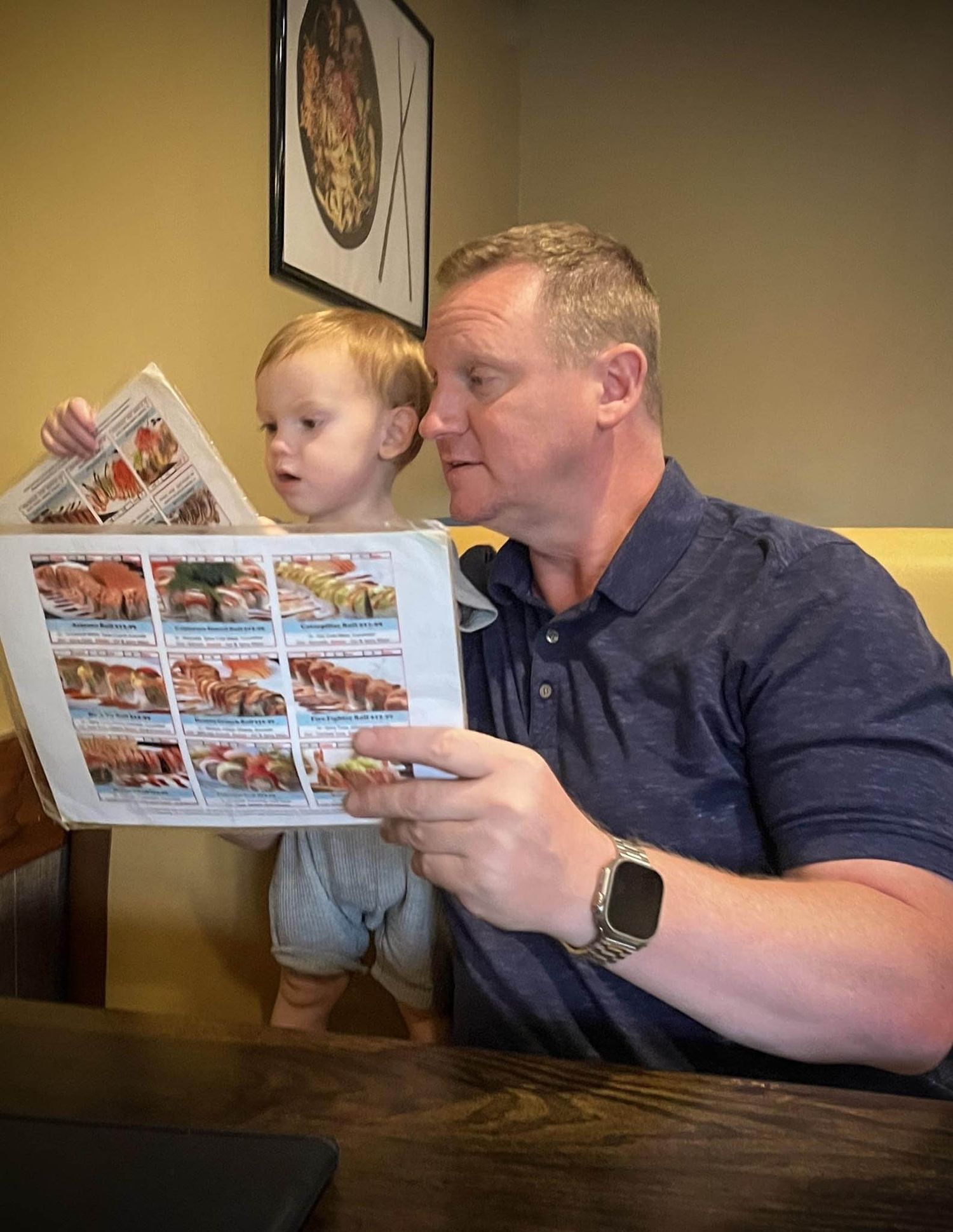 A person reads a photo album together with a young child while sitting at a wooden table indoors.