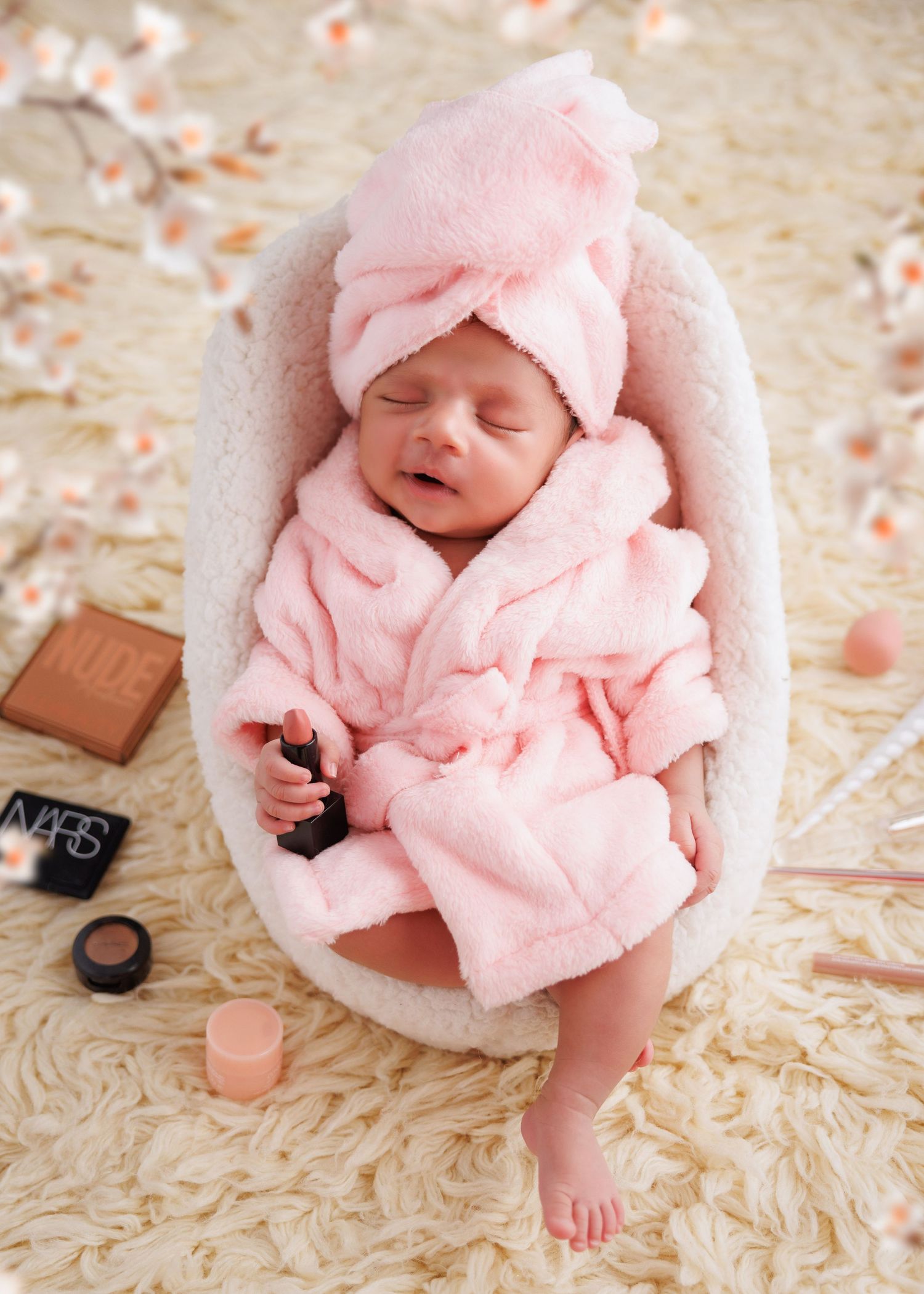 A sleeping baby in pink robe and towel lies in white cushioned bassinet with makeup items nearby.