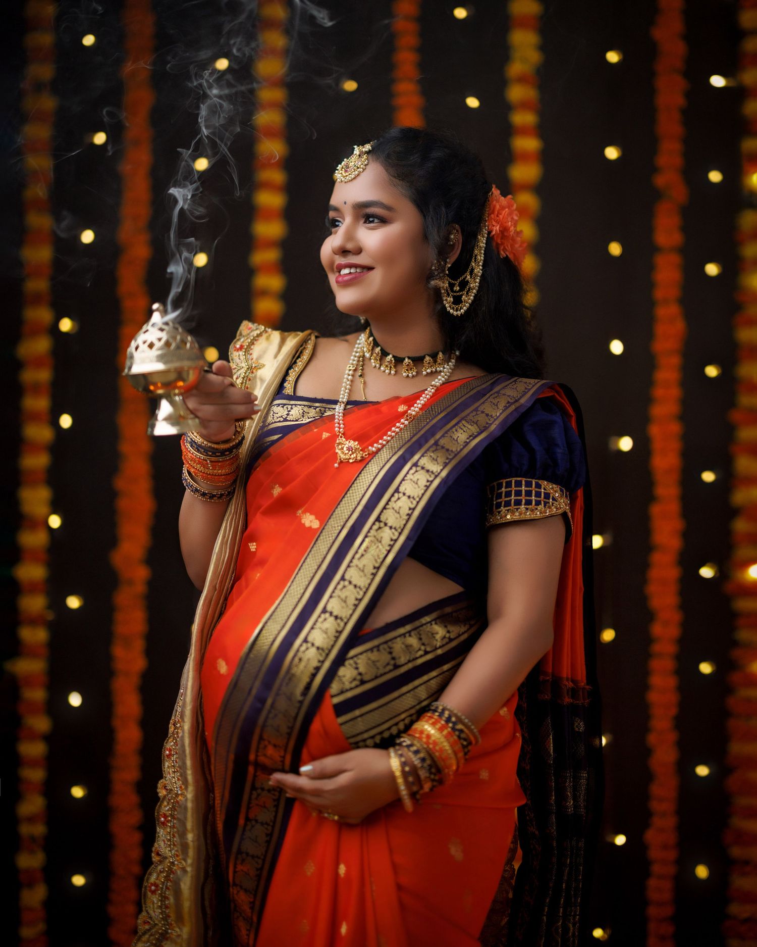A pregnant woman in traditional orange and navy Indian saree poses with a smoking incense holder against glowing lights.