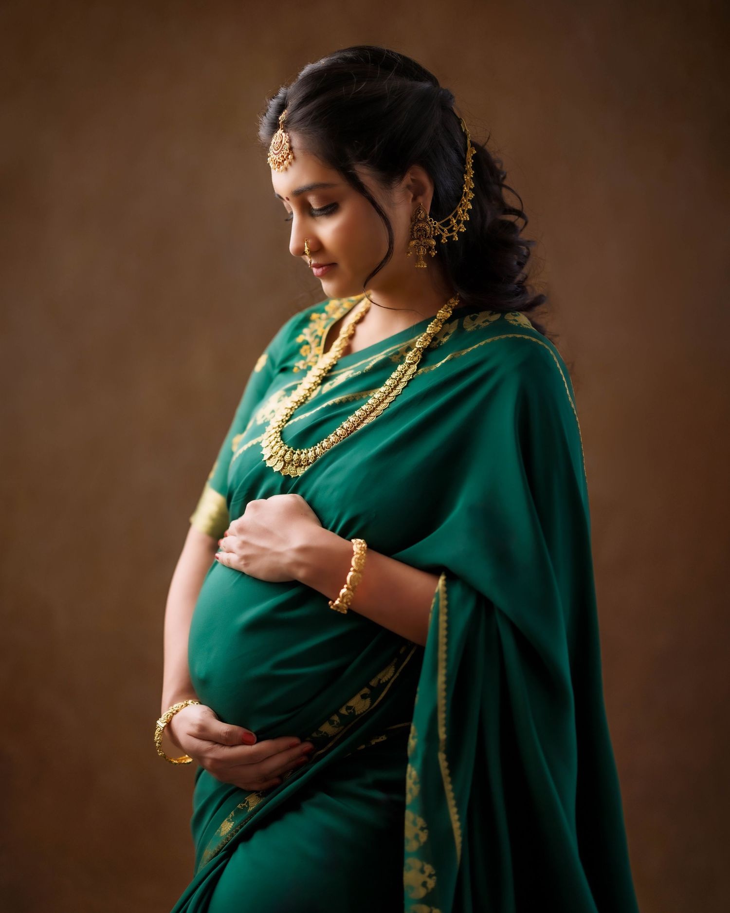 A person in a traditional emerald green saree with gold jewelry poses for a maternity photoshoot in a dark studio setting.