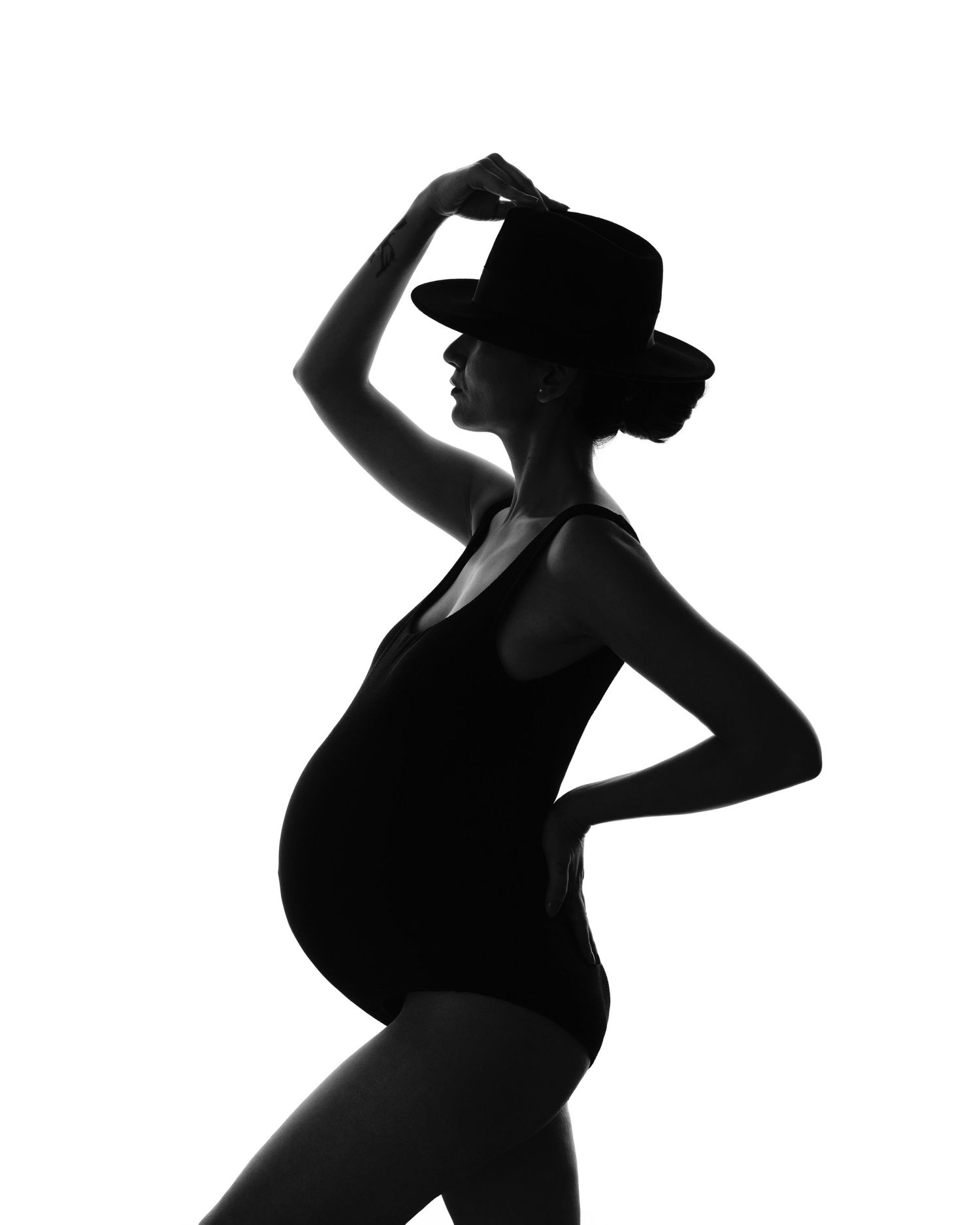Artistic black and white silhouette of a pregnant woman in a hat striking an elegant pose against white background.