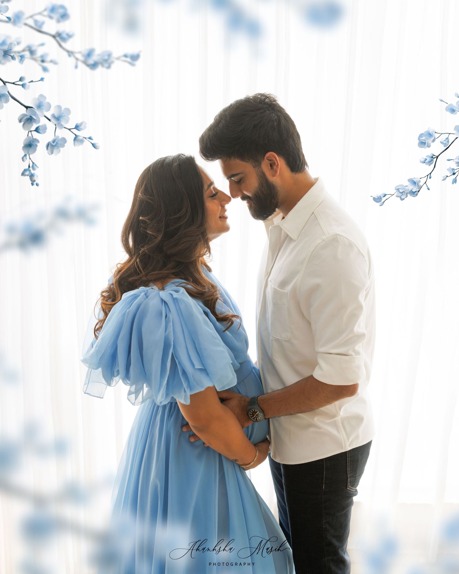 A romantic couple in a white shirt and blue dress embrace tenderly against a dreamy light background with blue floral accents.
