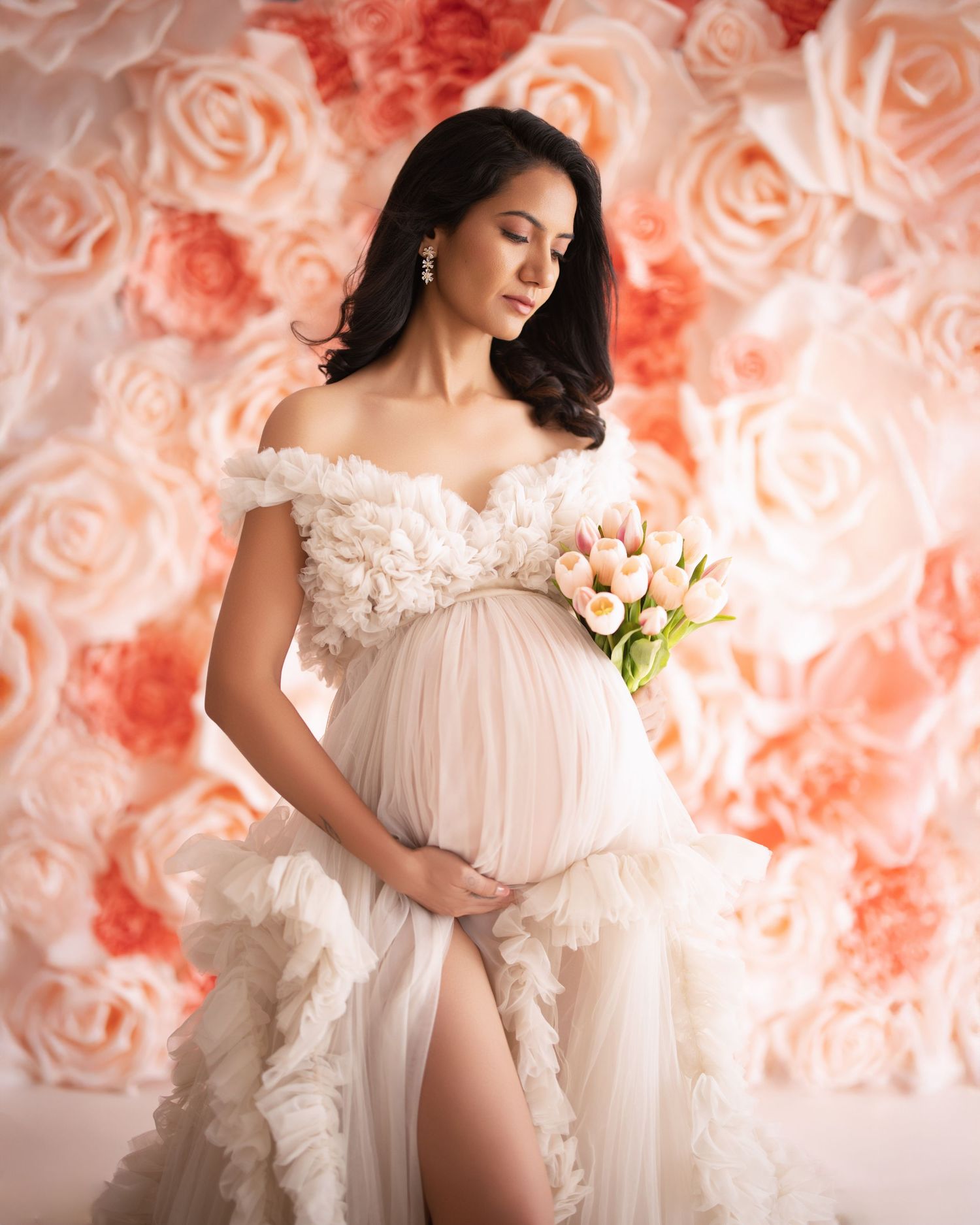 Elegant maternity portrait with off-shoulder white gown and pink roses backdrop, holding delicate tulips.