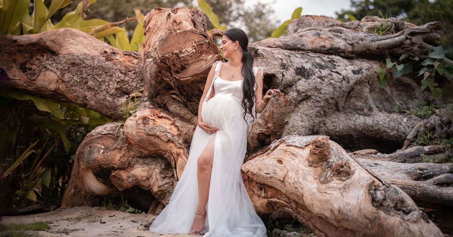 White maternity gown-clad figure posed gracefully against dramatic driftwood and natural scenery in outdoor setting.
