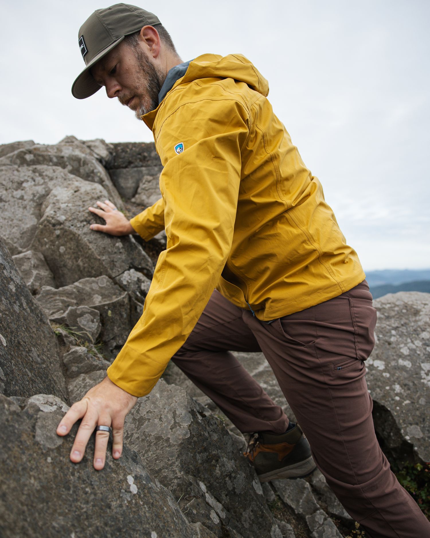 Rock climber in yellow rain jacket and cap carefully traverses rocky terrain on cloudy day.