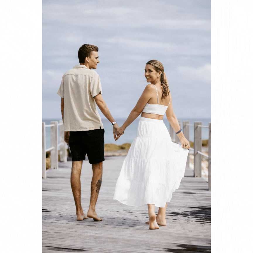 Casual couple holding hands while walking barefoot on wooden pier.