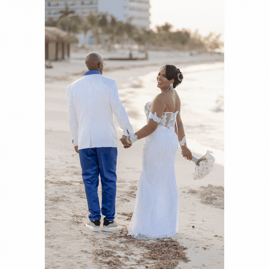 Newlywed couple walking on sandy beach with cityscape in background.