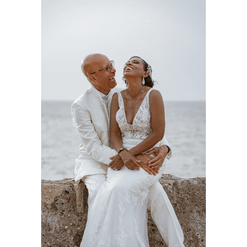 Joyful wedding couple sharing laughter on oceanfront wall.