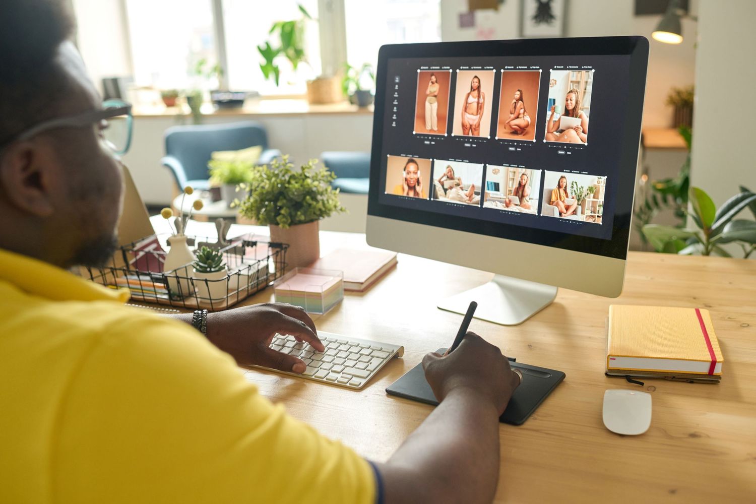 Person in yellow shirt working on digital illustrations at computer desk with plants nearby.
