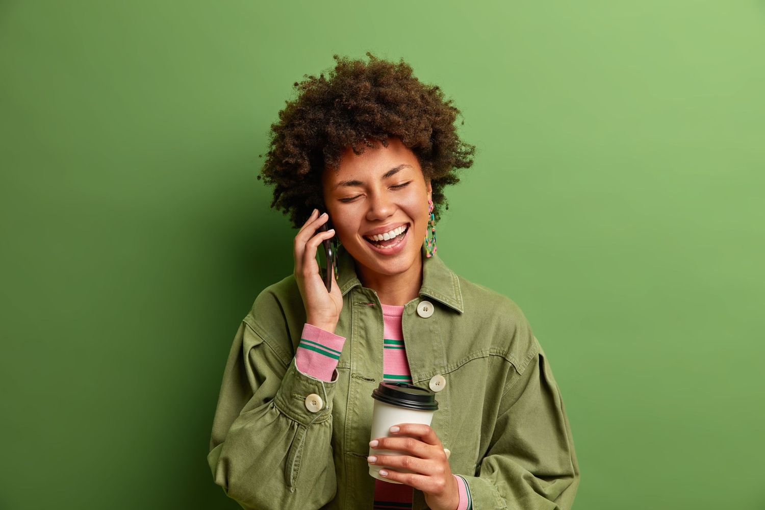 Person in olive jacket talking on phone and holding coffee against green background.