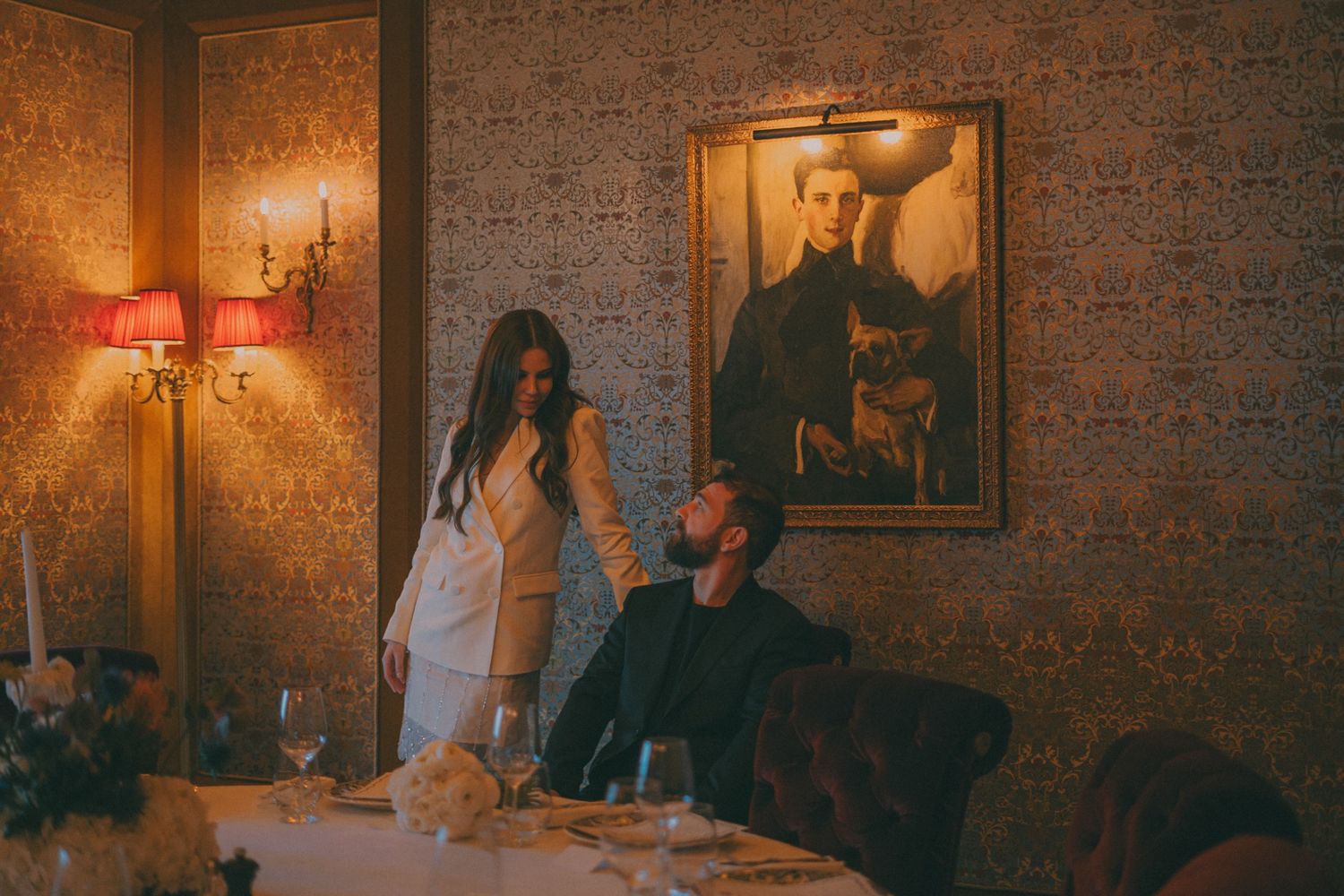 Photo de deux marié qui se regardent dans les yeux. Le marié est assis sur une chaise et la marié debout, la main sur le dossier