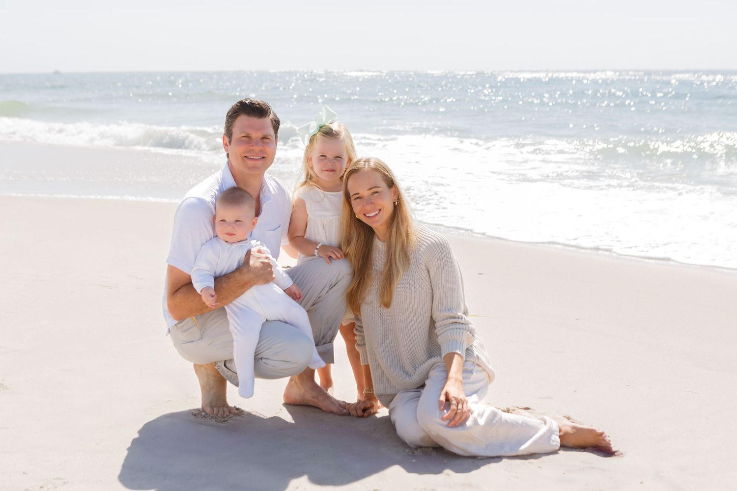 A family wearing white and beige clothing poses together on a sunny beach with ocean waves in the background.