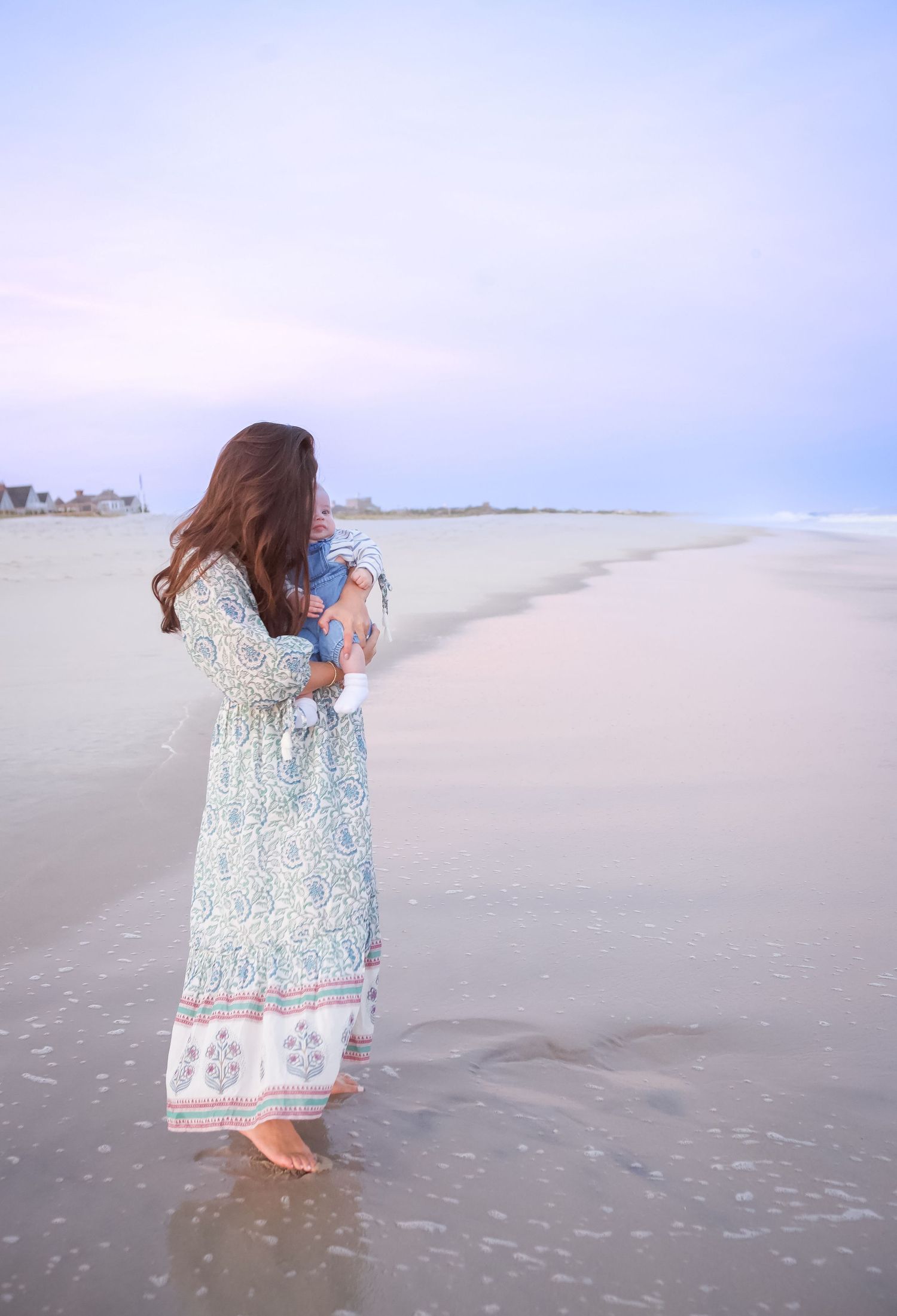 A mother in a floral dress kissing her baby along a misty Hamptons beach shoreline at dawn posing for photographer.
