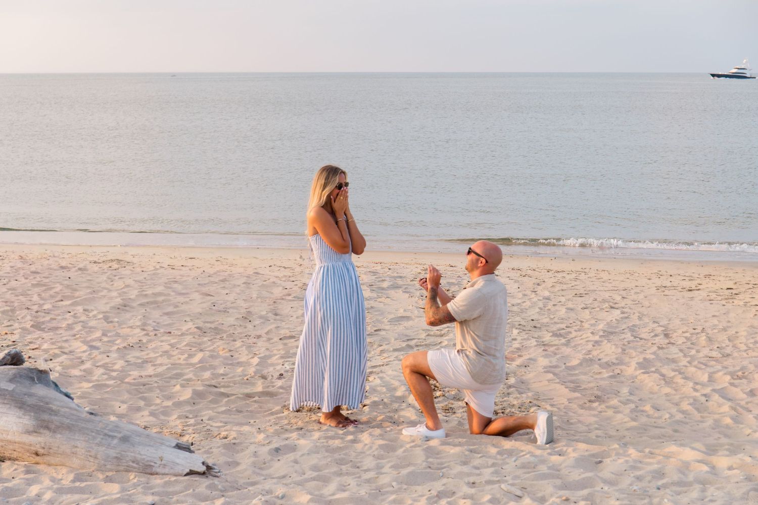 A romantic beach proposal at sunset with a couple sitting on white sand as waves roll in the background.