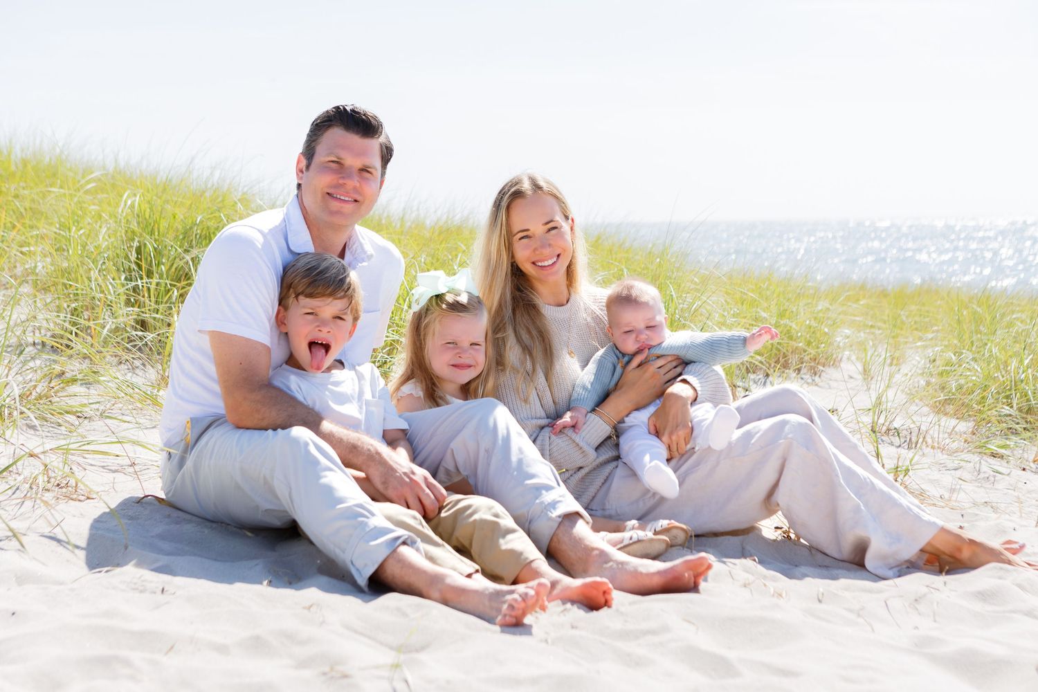 A group wearing white clothes relaxes together on a sunny beach with sand dunes in the background.