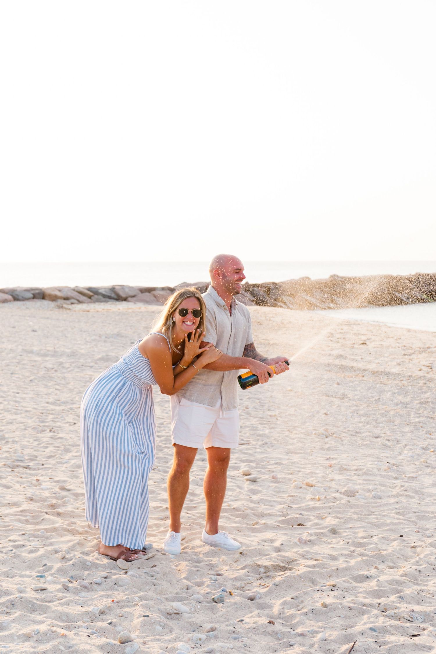 Two people share a joyful moment on a sandy beach during sunset, one bending over laughing while the other looks on.