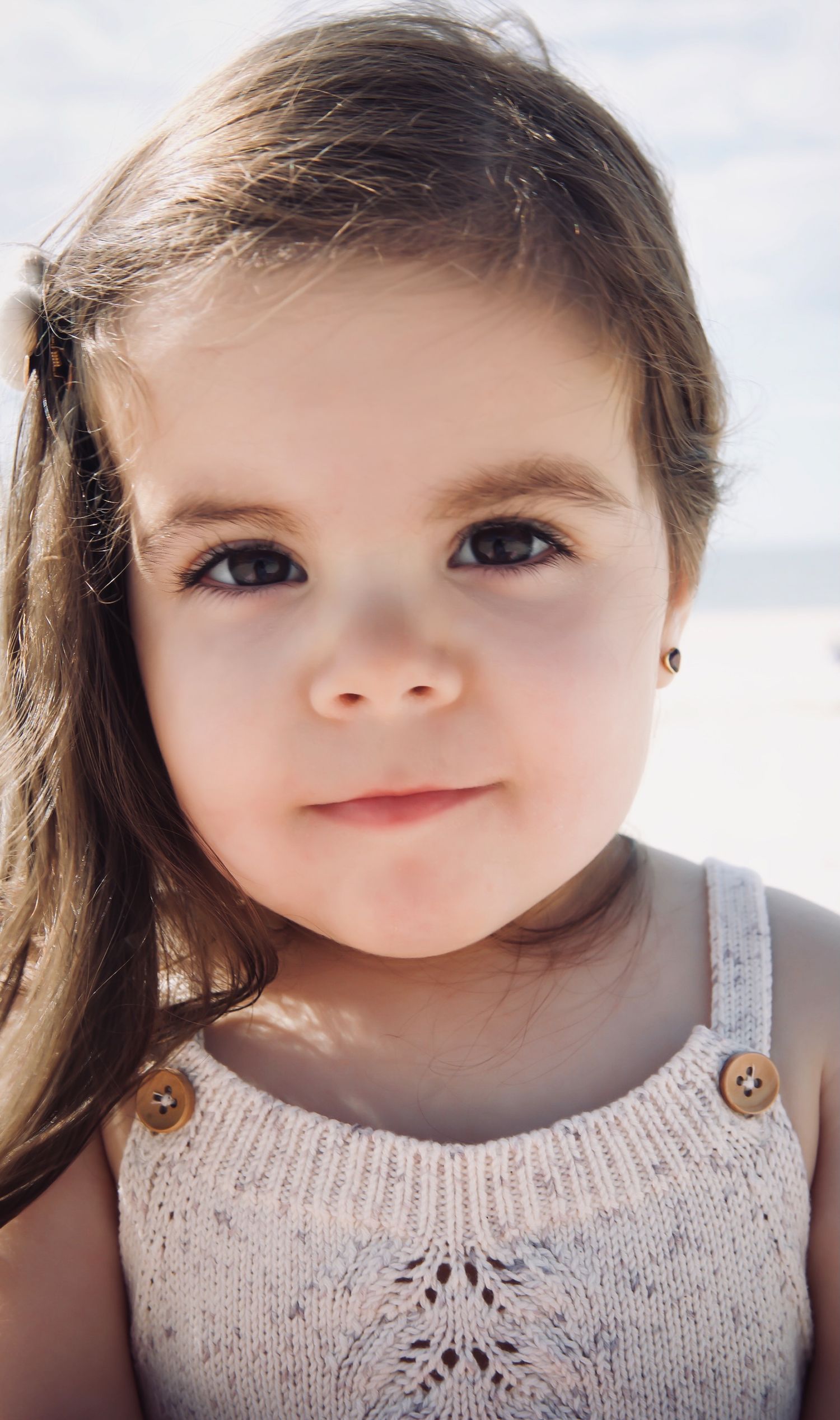 A young child in a knitted tank top with decorative buttons smiles softly at the camera in natural lighting at the beach in the Hamptons.