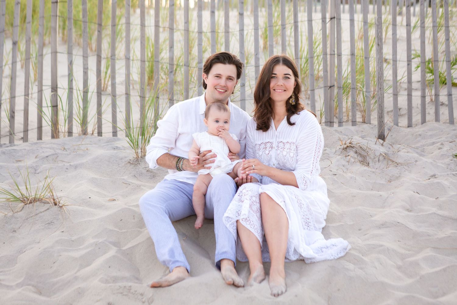 People in white clothing sitting together on a sandy beach with wooden fence posts in the background.