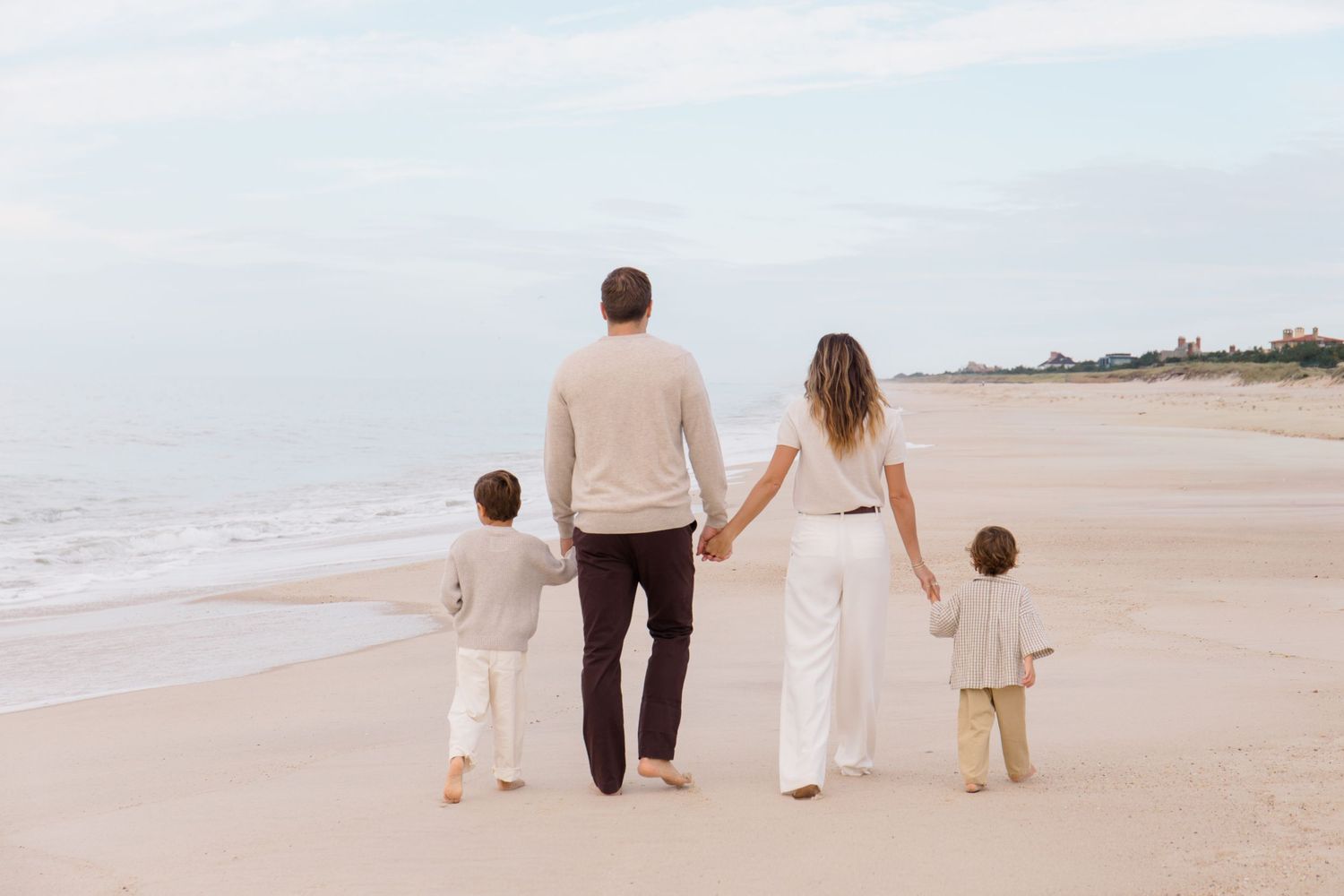 A family walks hand in hand along a serene beach shoreline on a cloudy day.
