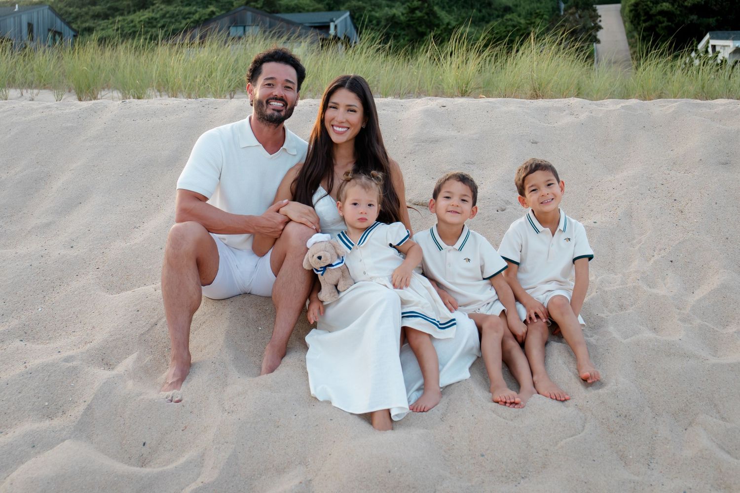 Family wearing white outfits sits together on sandy beach during sunset photoshoot.