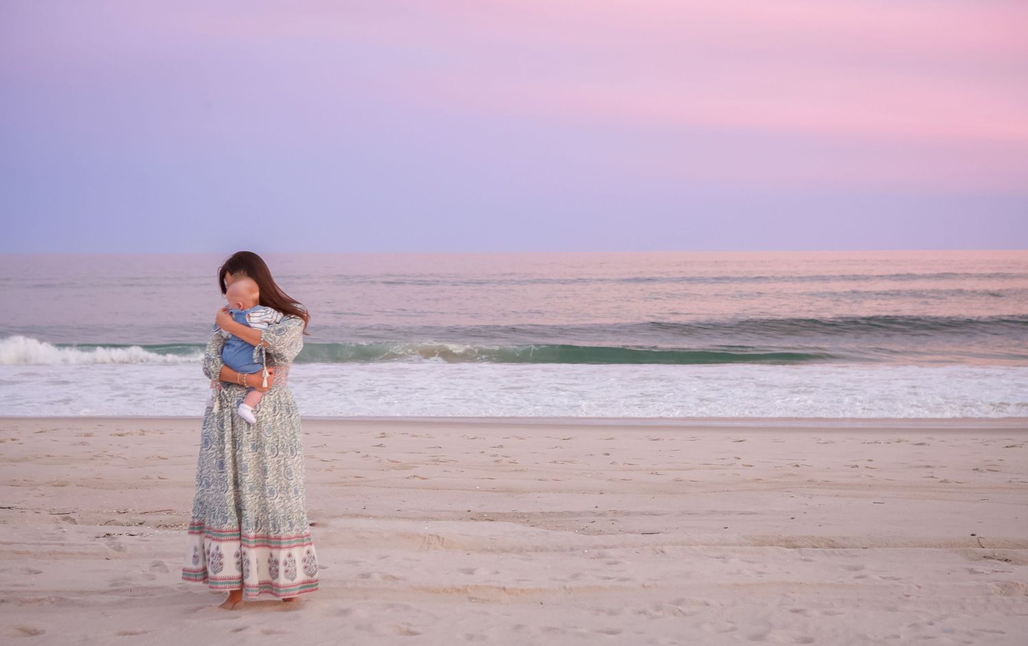 A figure in a flowing dress stands on a beach at sunset with pink and purple sky over the ocean waves.