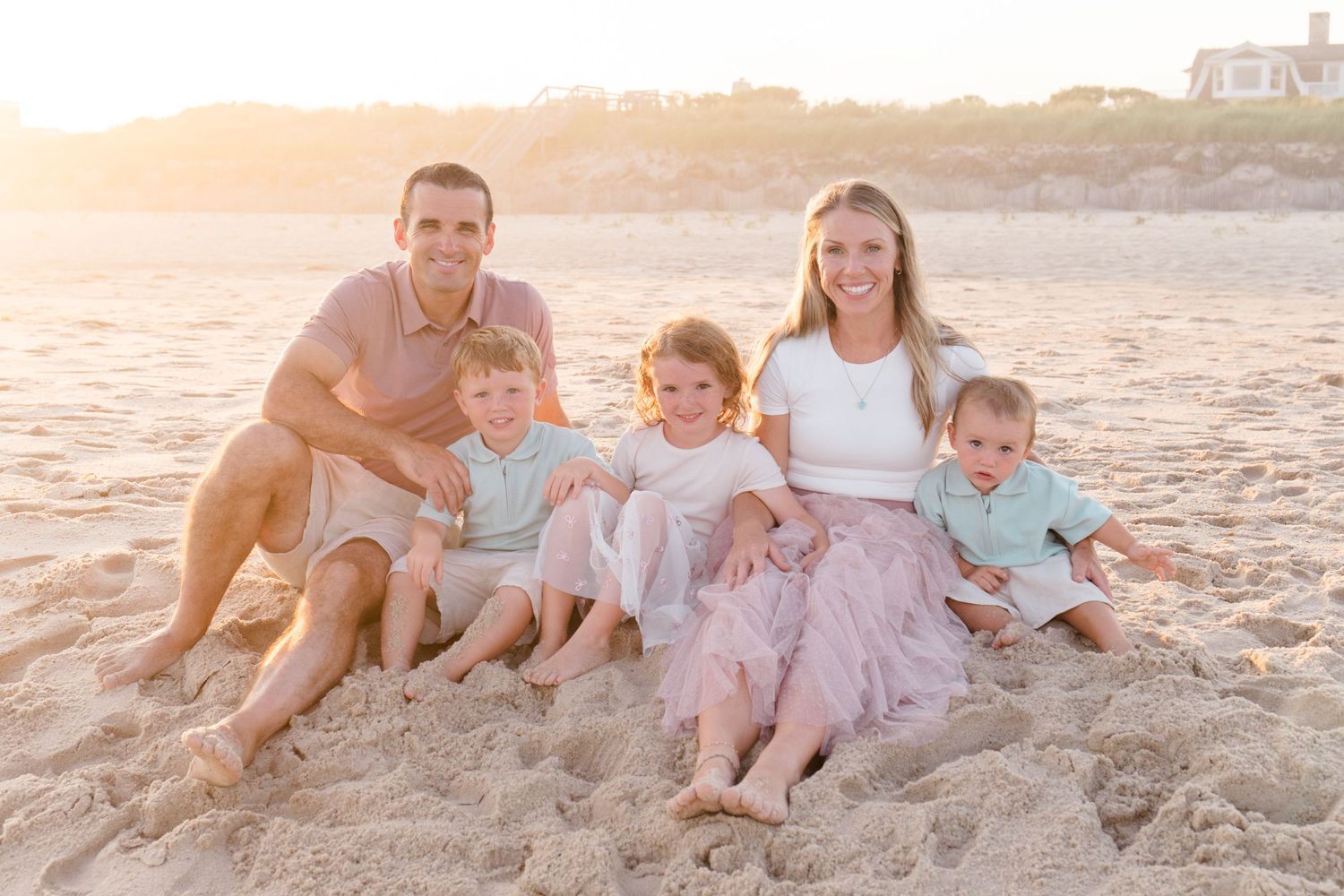 A family sits together on a sandy beach at sunset wearing coordinated pastel outfits for a photo session.