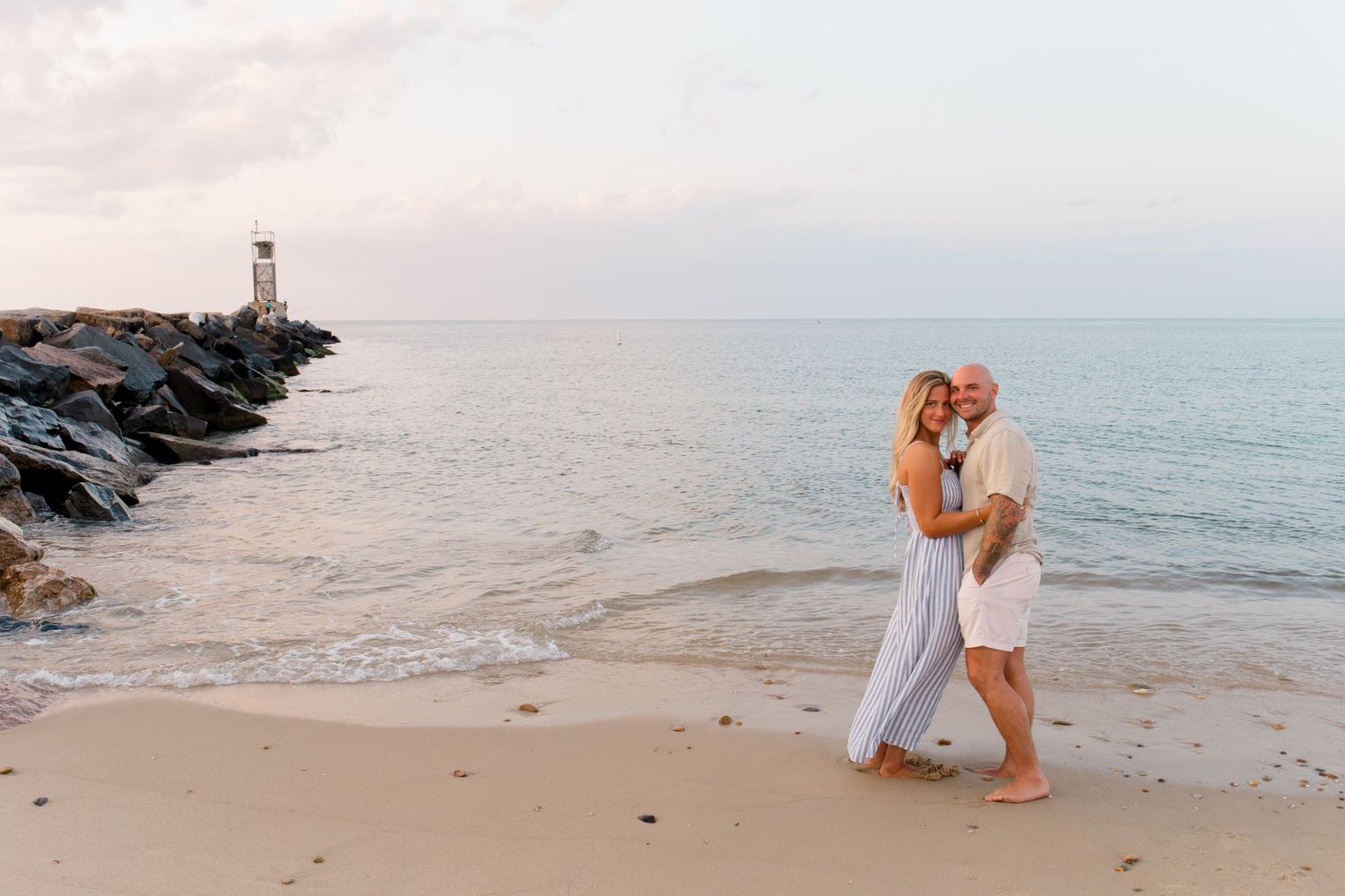 A couple embraces on a sandy Hamptons beach at sunset with a lighthouse on rocky jetty in the background posing for Hamptons photographer