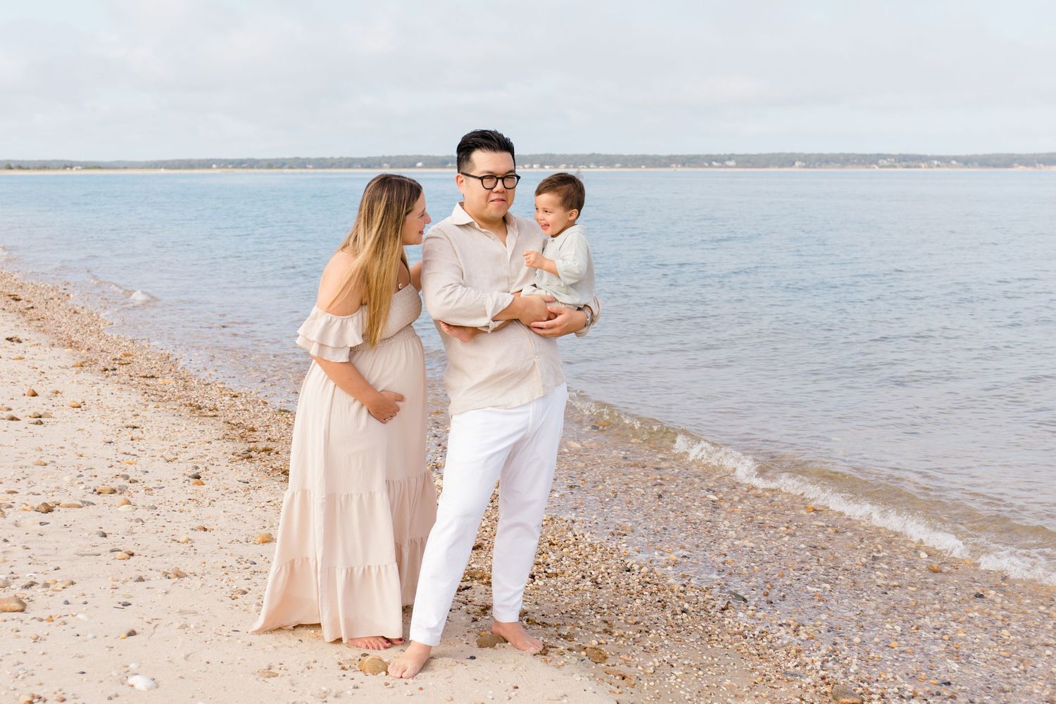 A family in light-colored outfits shares a tender moment while walking along a peaceful beach shoreline.
