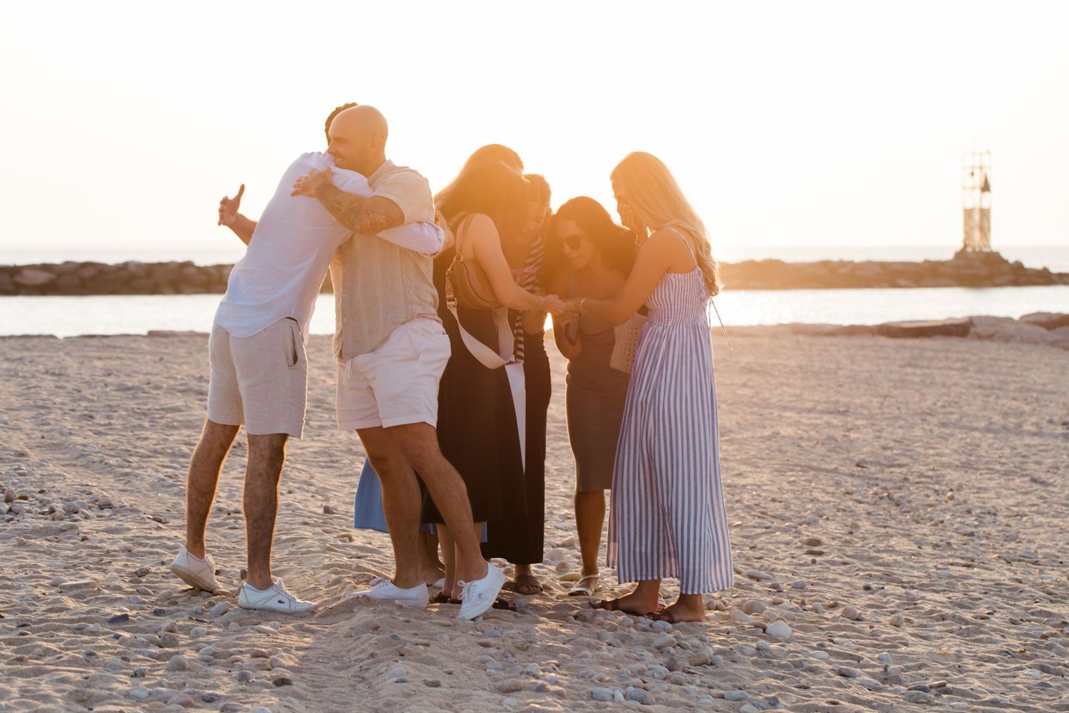 A group of friends embrace while standing on a beach at sunset with a lighthouse visible in the background.