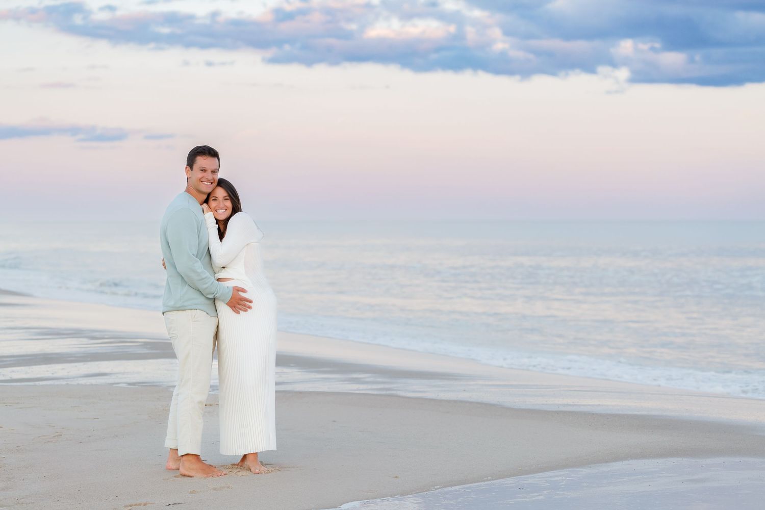 A pregnant couple in Sagaponack wearing white attire poses for photographer sharing a romantic moment on a serene Hamptons beach at sunset.