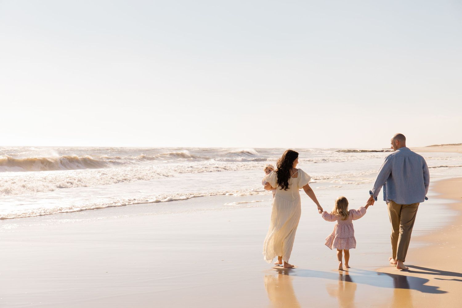 A family walks together along a sandy beach at sunset while holding hands and enjoying the ocean view.