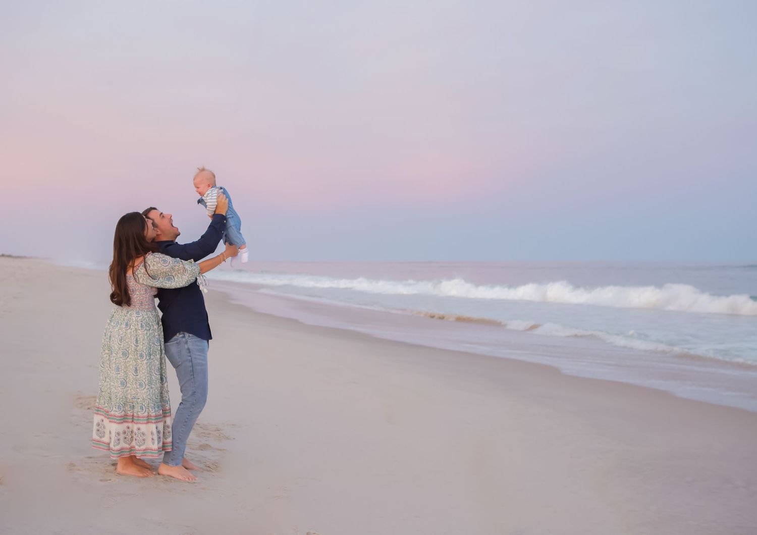 A father holds a baby up in the air on a peaceful Hamptons beach at sunset with gentle waves in the background.