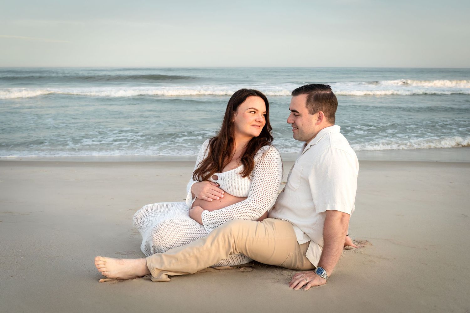 A couple sits together on a sandy beach at sunset with ocean waves rolling in the background.