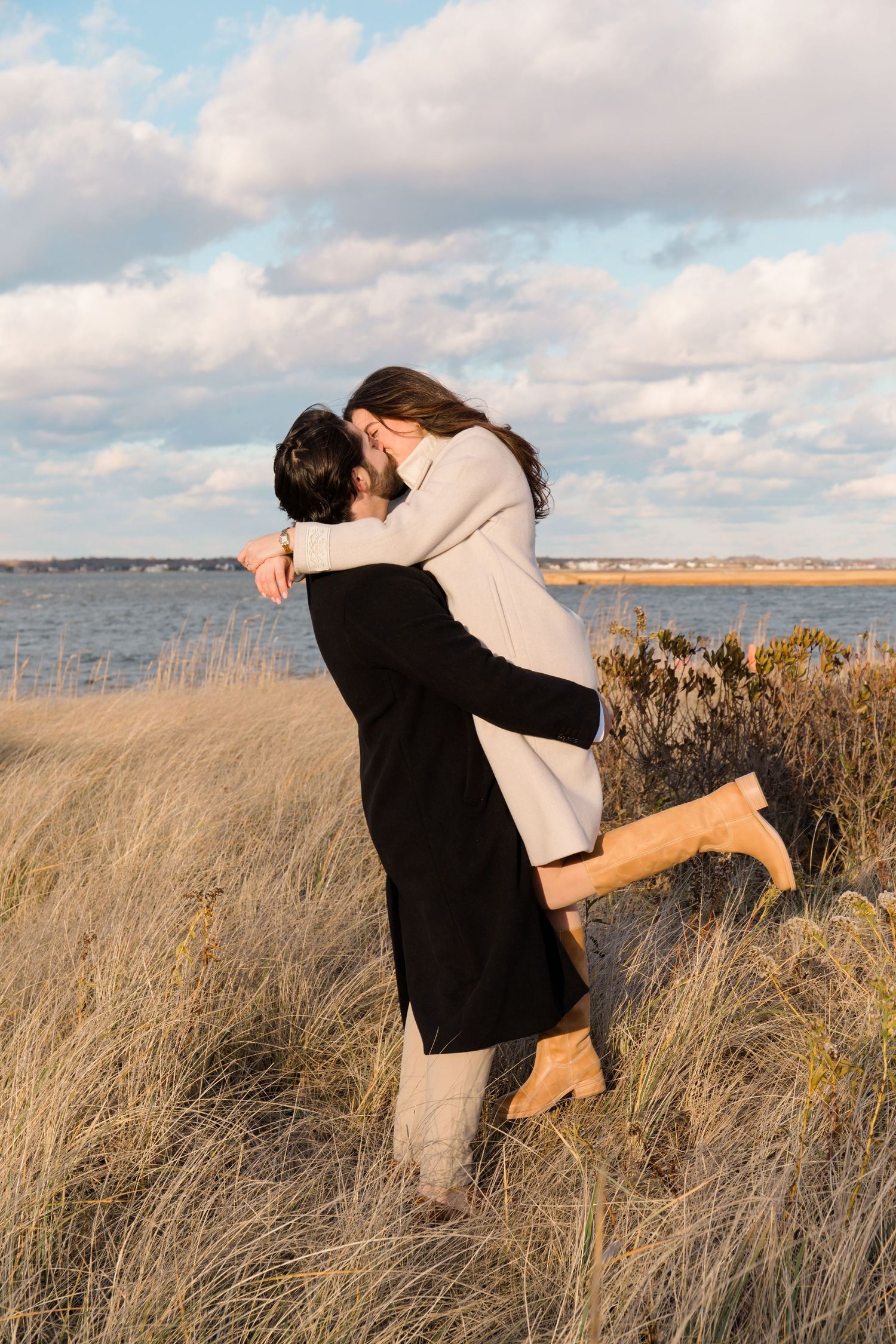 Couple embracing in tall grass by the ocean in Hampton Bays during sunset with cloudy skies posing for a photographer