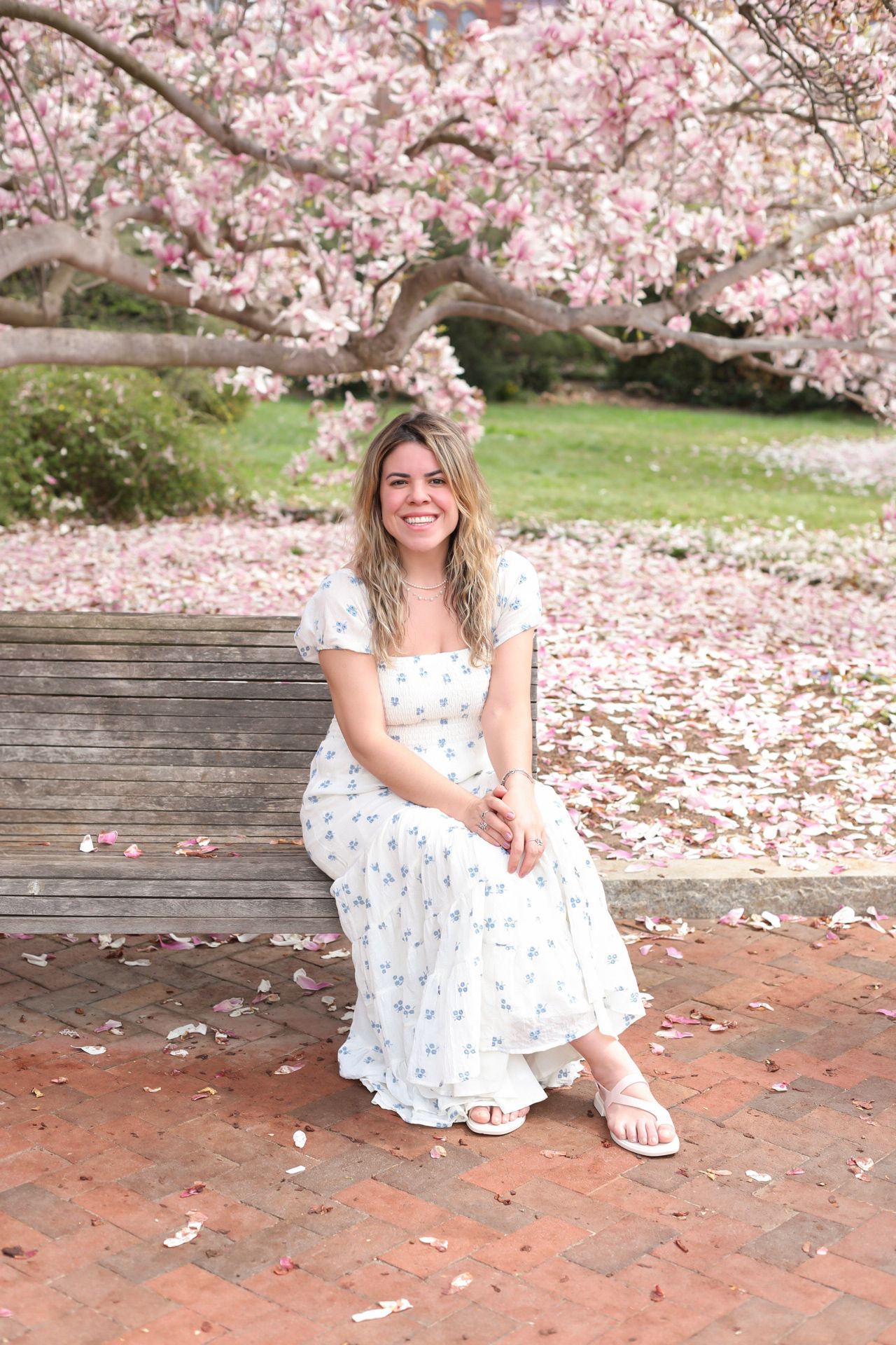 A photographer in a white floral dress sits on brick pavement under blooming cherry blossom trees in spring.