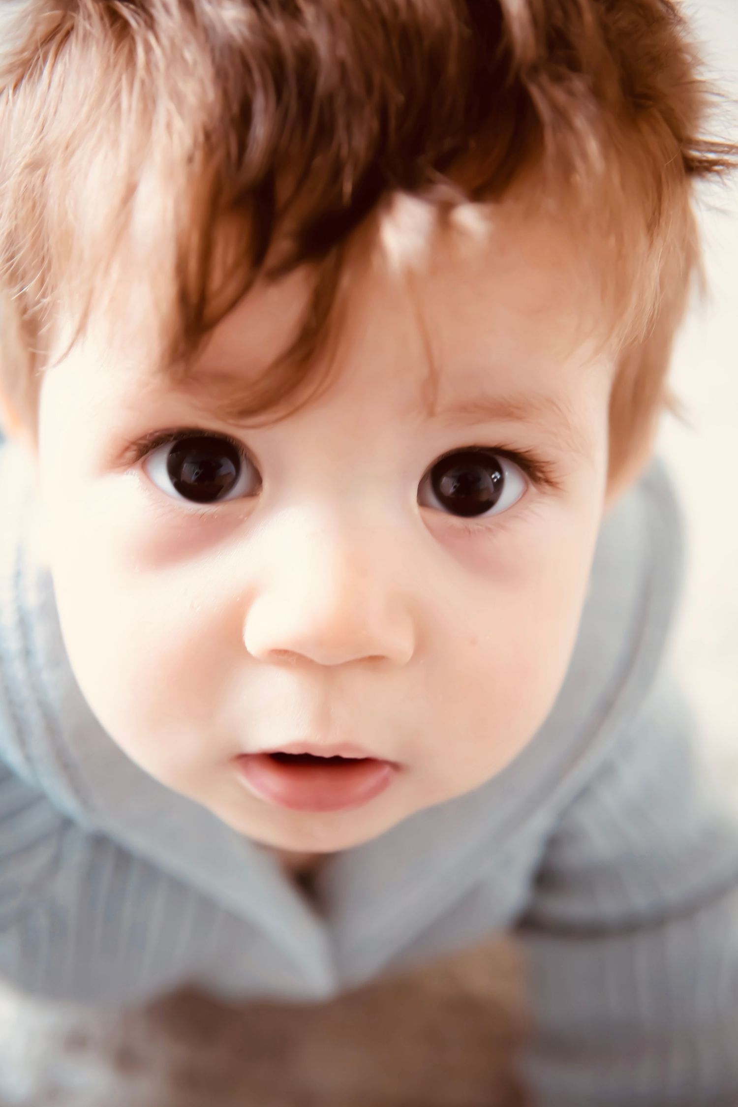 Close up portrait of a baby with brown hair and big brown eyes wearing a light gray top.