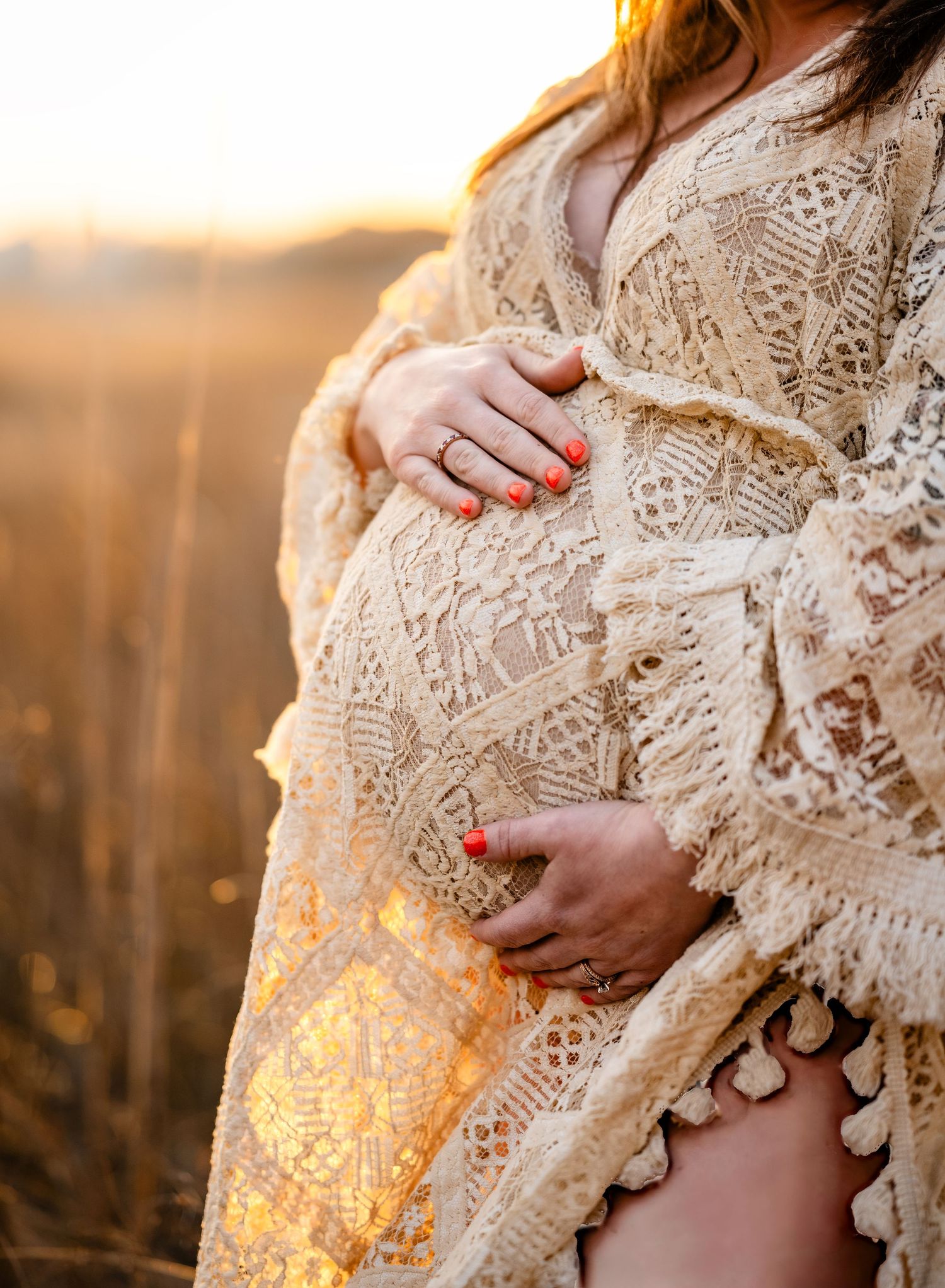 Pregnant woman in lace dress cradles baby bump during golden sunset in field.