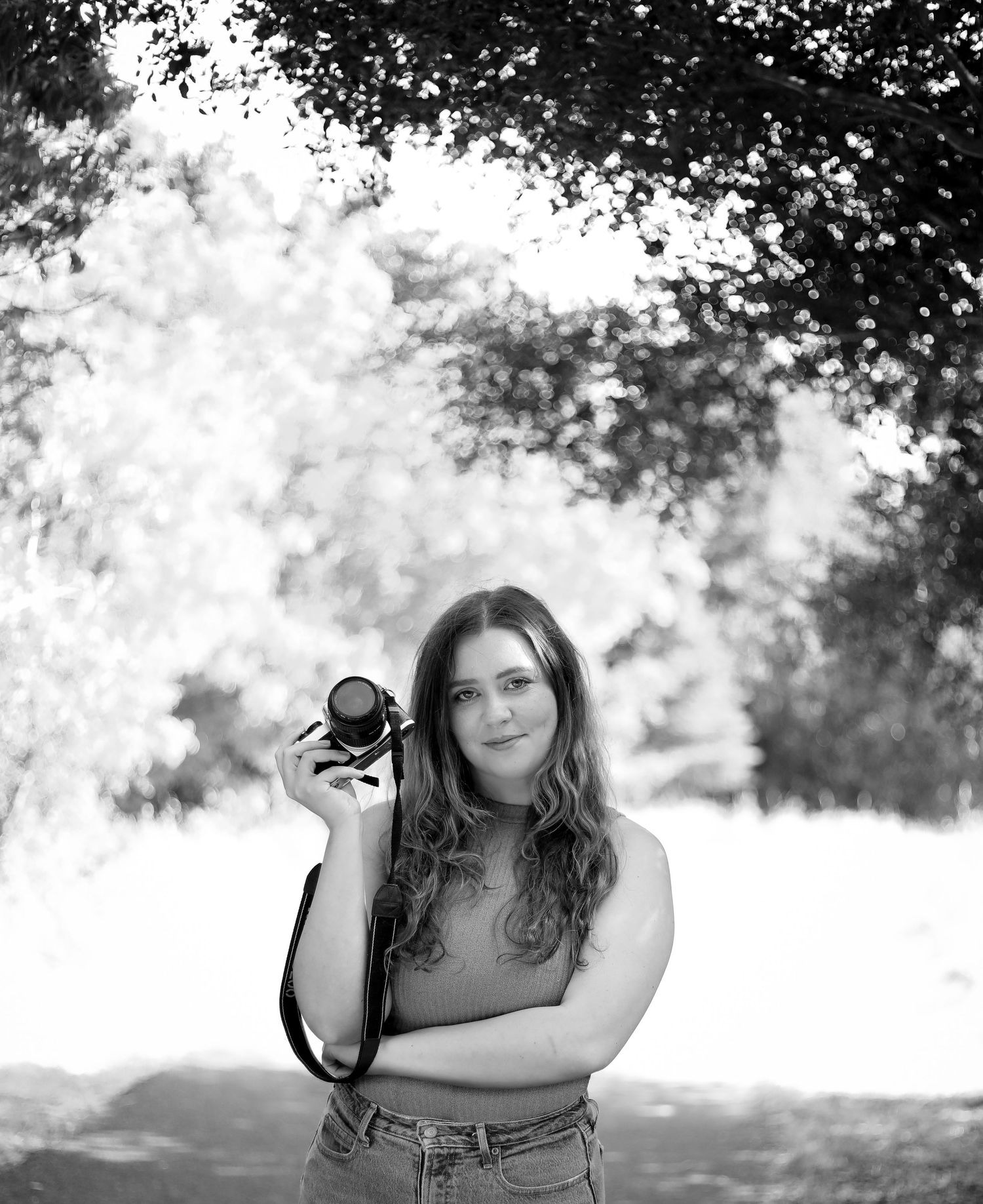 A black and white photograph shows a person in casual attire holding a camera outdoors with trees in the background.