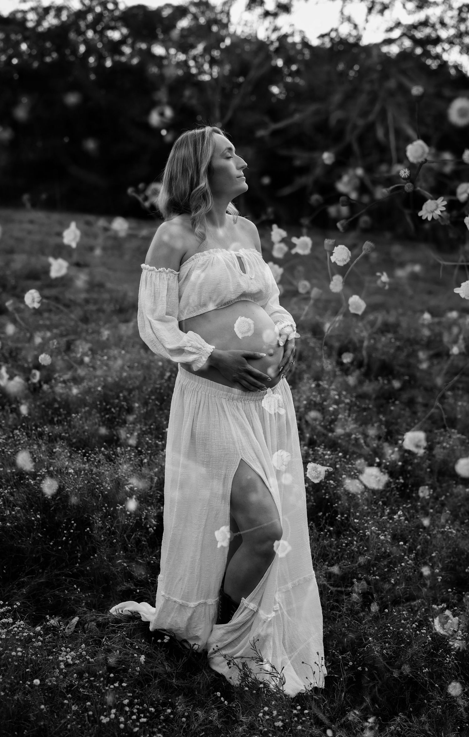 Black and white maternity photo of pregnant woman in white off-shoulder dress holding belly in a flower field.