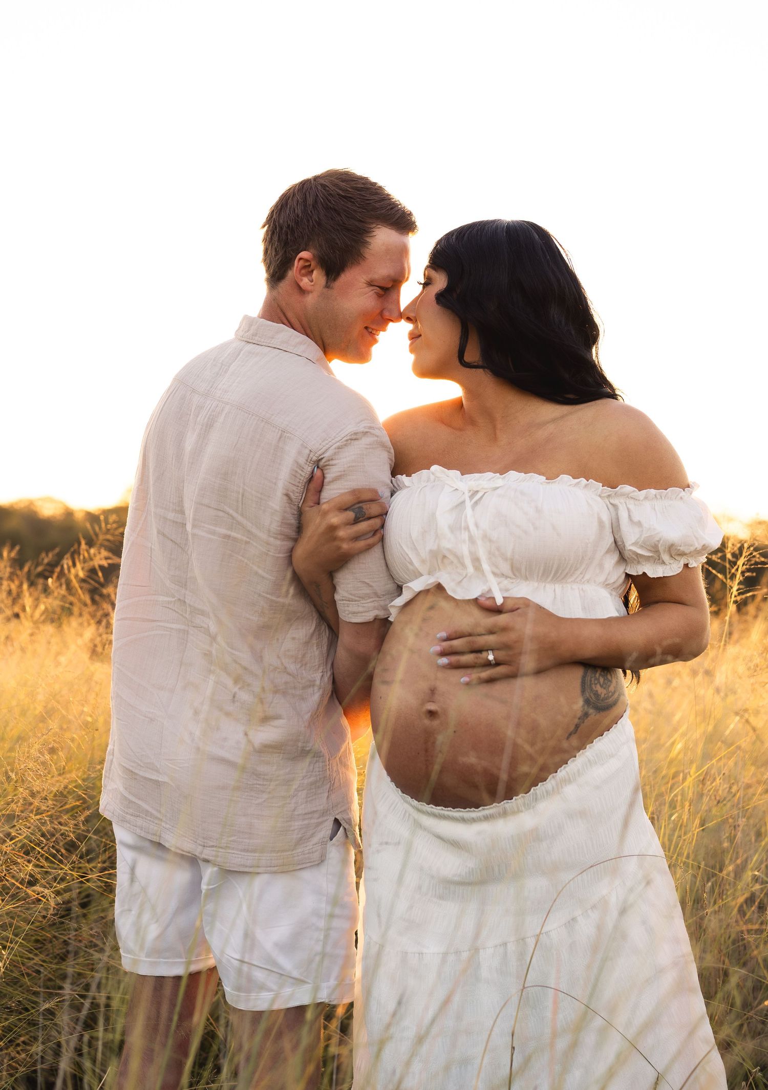 Expecting couple almost kissing in sun-drenched grass field, woman in white off-shoulder maternity dress showing baby bump.
