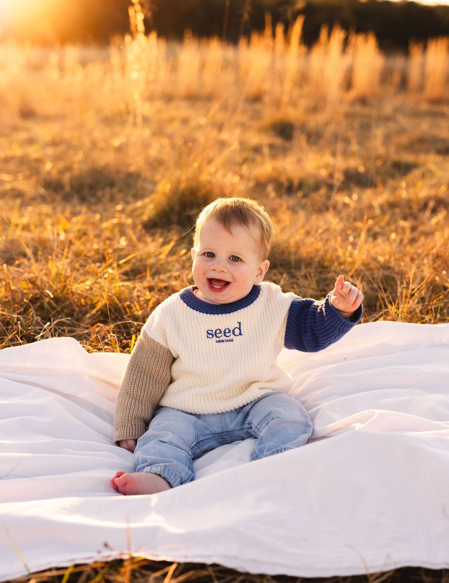 Happy baby boy sitting on white blanket in golden field wearing a cream and navy Seed sweater with jeans.