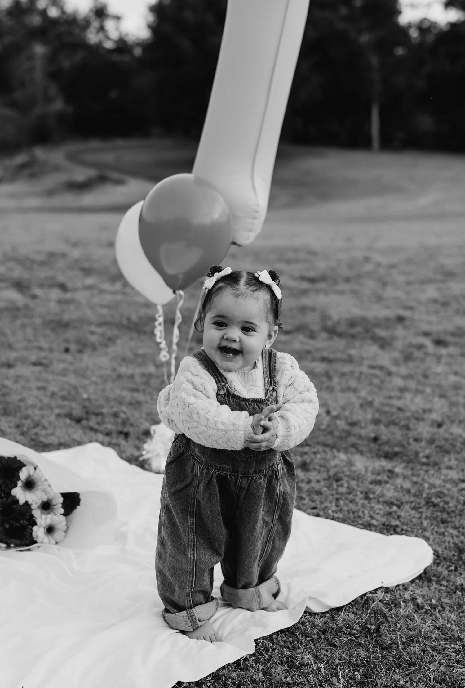 A smiling baby girl in overalls and a bow sits on a white blanket outdoors with balloons at a first birthday celebration.