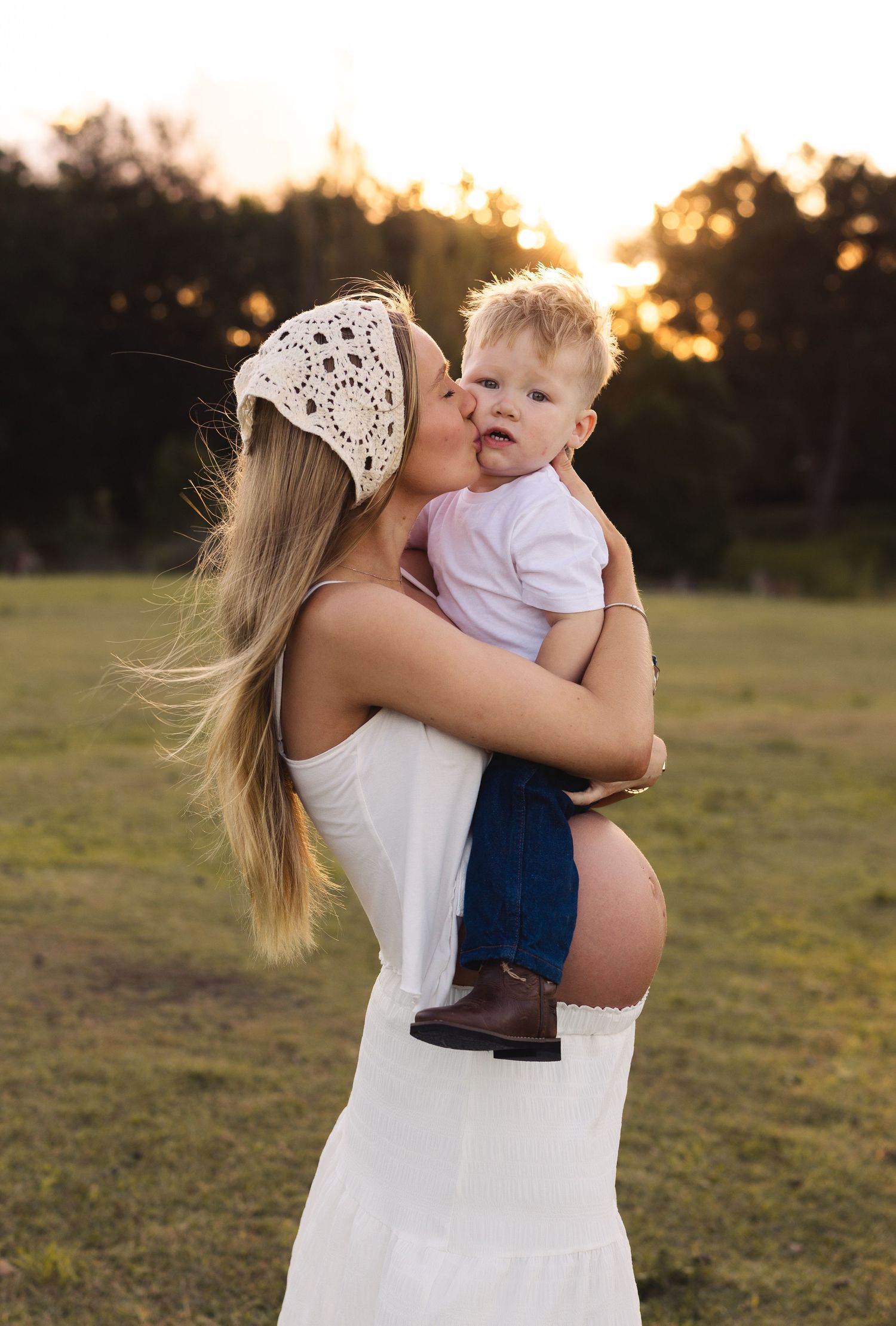 Pregnant woman in white dress kissing toddler boy at golden hour in a green field.