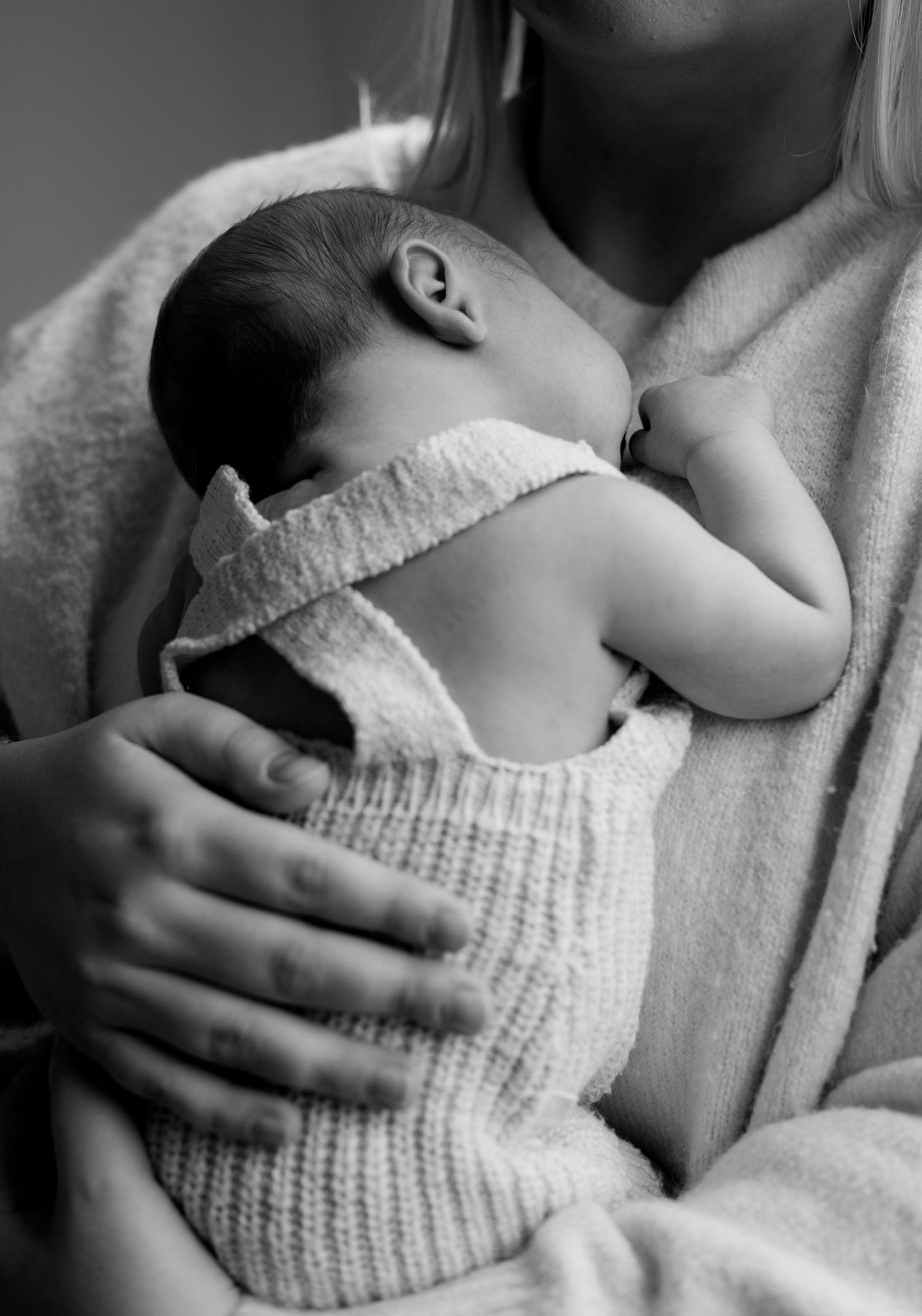 Black and white photo of mother cradling sleeping newborn baby wrapped in knit blanket.