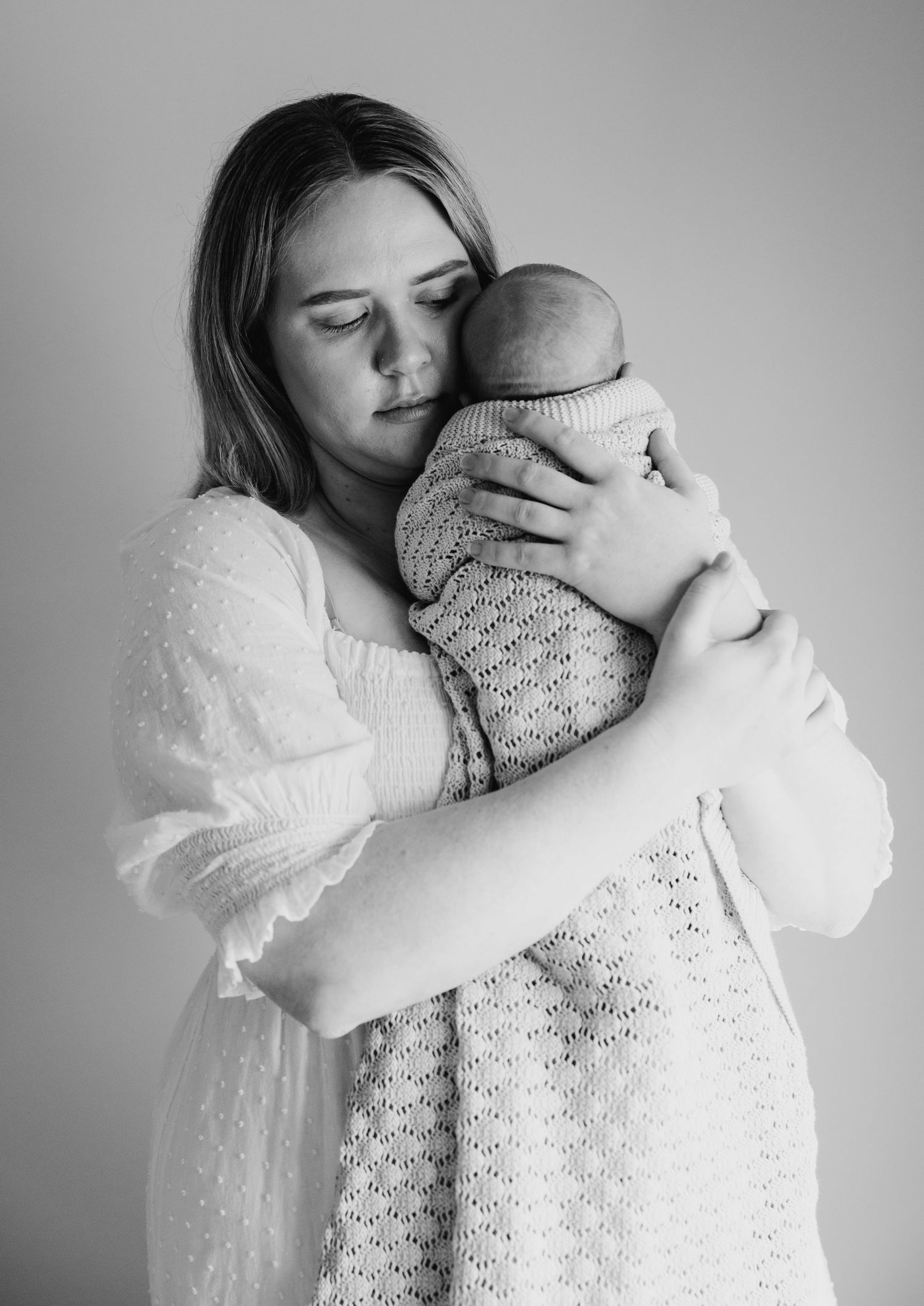 Emotional mother in white dress tenderly holds sleeping newborn close in B&W.